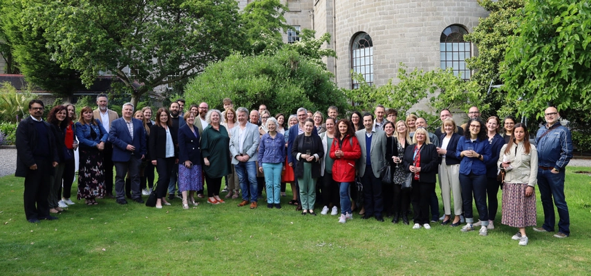 Large group of people in the Provost's garden, all smiling at the camera. They are standing on grass and there are trees and a tall stone building in the background.
