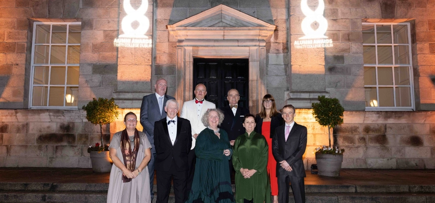 * Group of 9 people standing on the steps of the Dining Hall. Photo taken at night time, with projections of the Trinity Alumni Awards on the wall in the background.