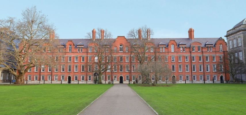 Image of the facade of the Rubrics in Trinity College Dublin. A four storey, red bricked building with trees, grass and a straight path in the foreground.