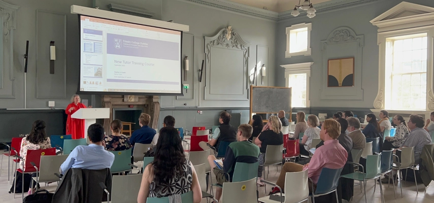 Room with seated audience listening to the Provost speak. There is a PowerPoint presentation behind the provost reading ‘New Tutor Training Course’.