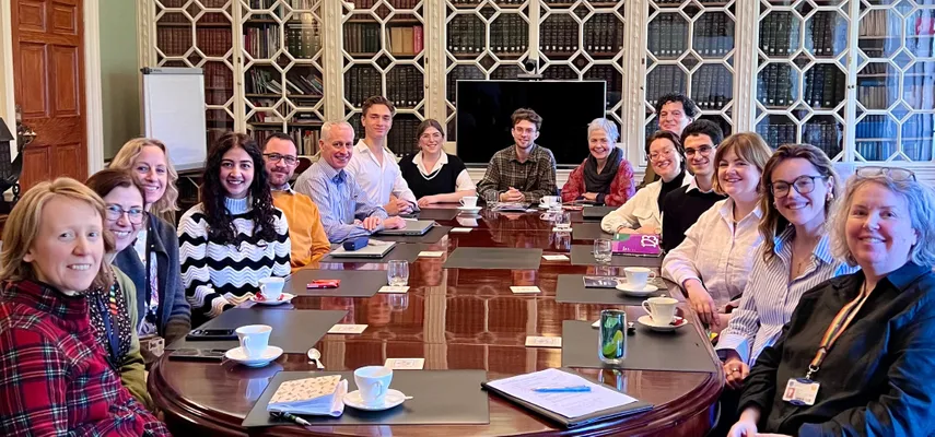 Group of 16 people sitting around a large, brown table. They are all smiling and turned towards the camera.