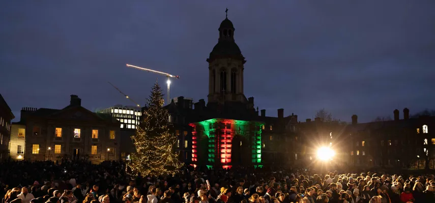 Large crowd gathered in Front Square at night time. Lit up Christmas Tree and Campanile lit up in red and green in the centre of photograph.