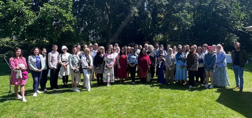 Large group photo taken in a garden. The crowd are standing on grass, with trees in the background and the sun is shining.