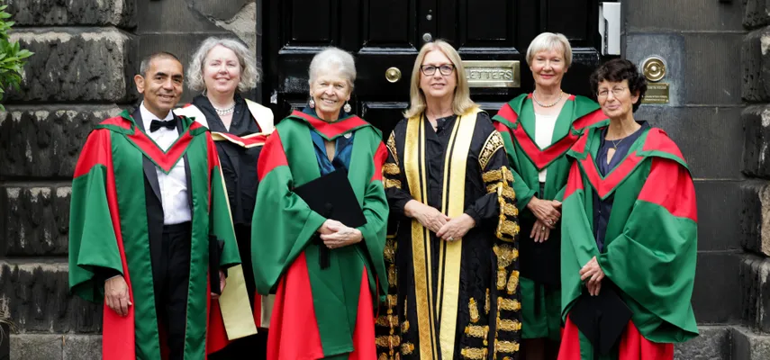 Six people standing on stone steps in front of a black door. They are all wearing ceremonial academic gowns.