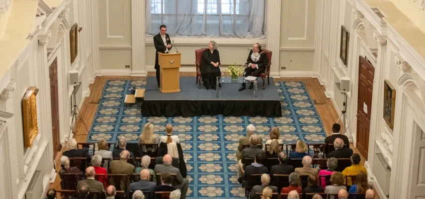 Photo looking down on an event with audience sitting in front of a small stage, with 3 people on it. Two of the people on stage are sitting, and one is standing.