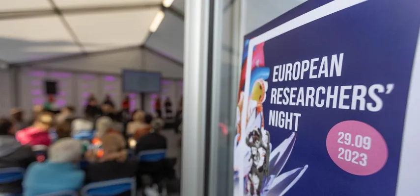 Photo taken looking in the door of a tent with people sitting, facing the back. European Researchers' Night poster is stuck to the door, to the right of the image.