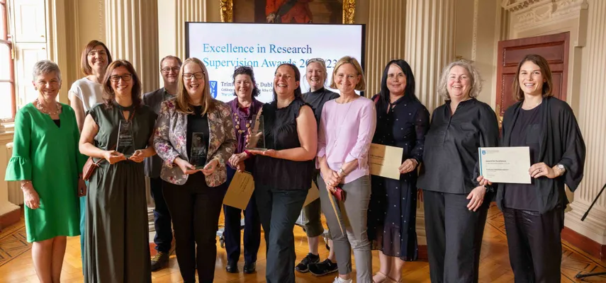 Group of people standing and smiling for a photo. Some are holding certificates, others are holding glass awards. Screen in the background reading 'Excellence in Research Supervision Awards 2022/2023'.