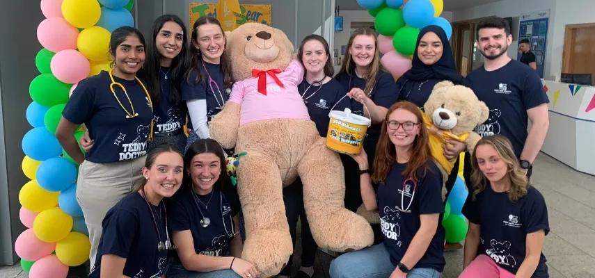 Group of students gathered round a large teddy bear. Colourful baloon arch in backgrond.