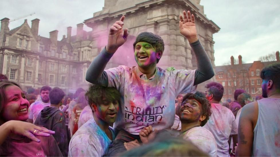A trio of students from TCD Indian Soc celebrating in a crowd, covered in colourful powder