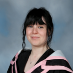 Adjunct Tara Kerins Aylmer wearing black and pink academic dress against a grey and blue studio background.