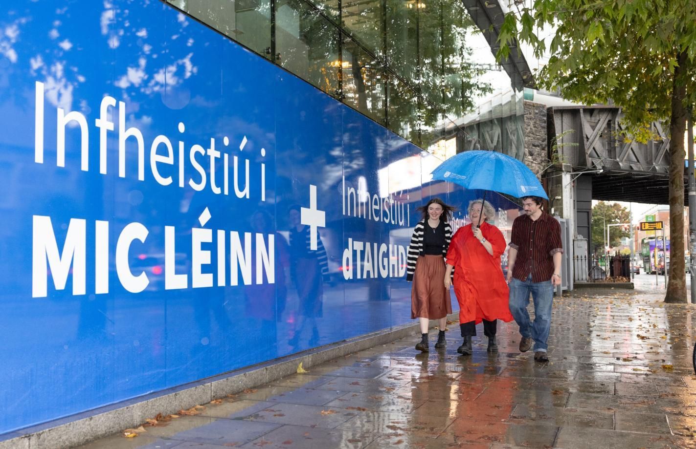 Three people walk together along a large Irish-language sign