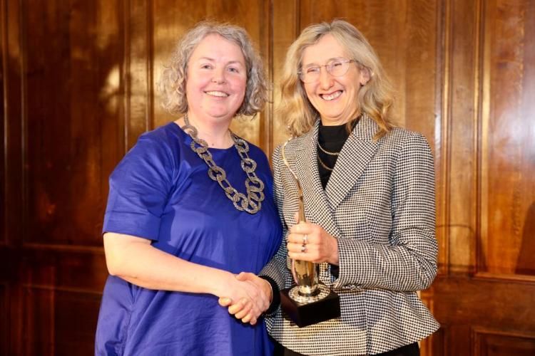 Provost Linda Doyle in a blue dress gives the Provost Innovation Award to Professor Orla Hardiman, who is wearing a black and white dress.