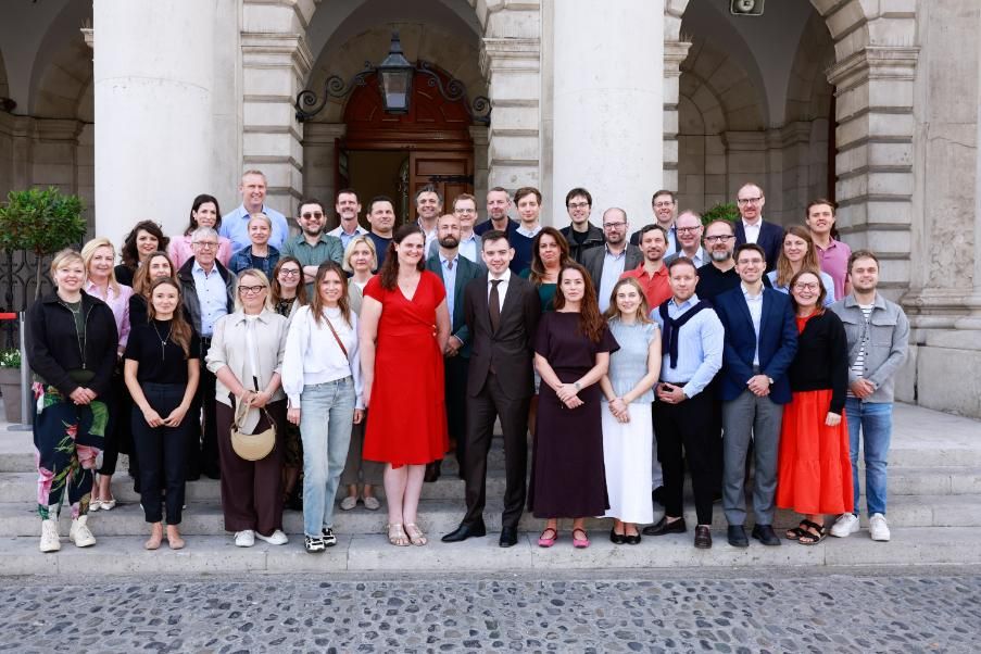 The large group of Research Attachés stand for a group photo on the steps of Trinity's Public Theatre. The columns and arches of the building are visible in the background.