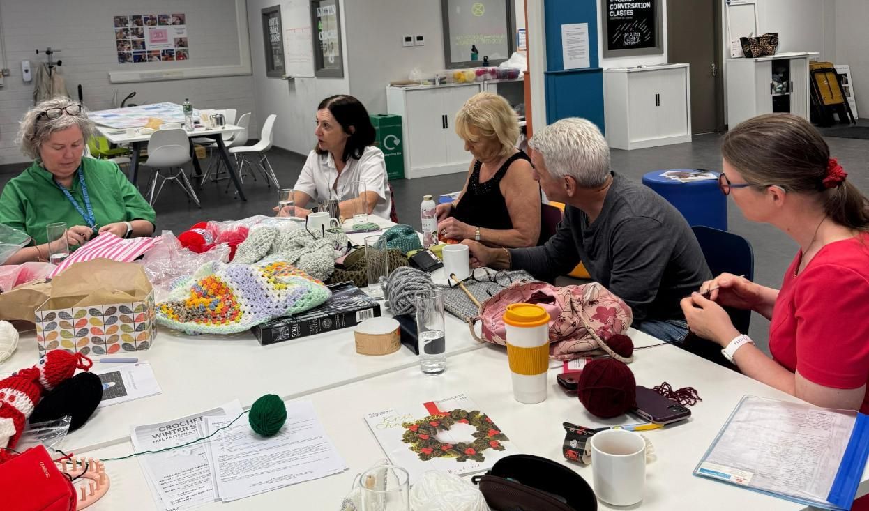 The Crafty Creatures group gathered around a large table, working on their crochet projects. There is yarn, instruction sheets and crochet projects on the table. Linda is seated at the table to the far left of the frame.
