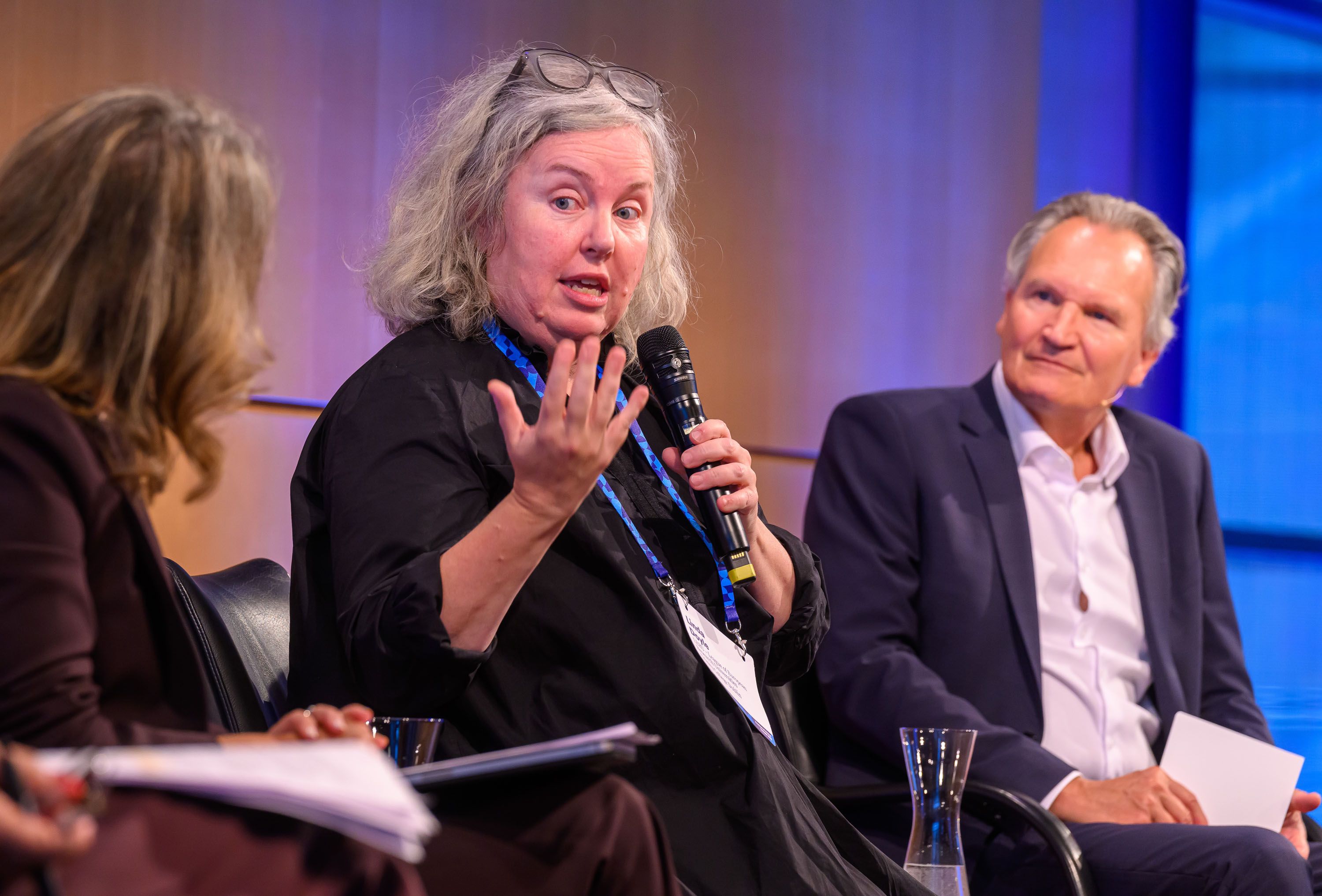 Close-up of Linda speaking on stage at the Hamburg Science Summit. She is speaking into a mic and gesturing towards the panelist to her right, who is mostly out of frame. The panel MC is visible, seated to her left.