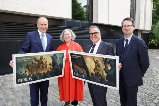Taoiseach Micheál Martin, Provost Linda Doyle, David McRedmond (An Post), and Prof. Patrick Geoghegan standing for a photo. They are holding two large stamps depicting illustrations of Daniel O'Connell speaking to crowds of people.