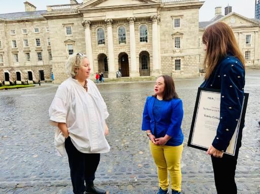 Provost Linda Doyle, Dr. Esther Murphy and Ms. Mei Lin Yap - Front Square at Trinity College Dublin, talking and smiling. Esther wears a navy jacket with gold bee details and holds a framed certificate. Mei Lin Yap wears blue and yellow clothing, and the Provost Linda Doyle wears a white blouse. The cobblestone ground and historic stone buildings frame the scene.