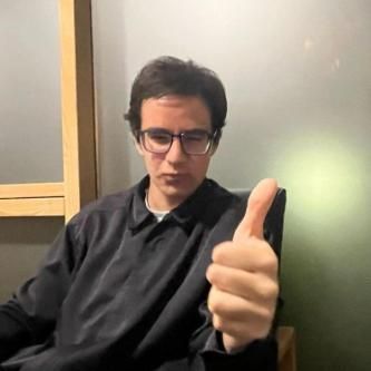 A student with brown hair pursing their lips at the camera. He has his thumb up.