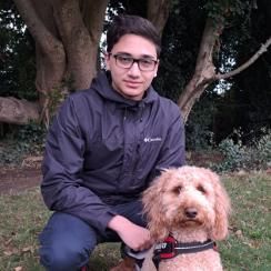 Student with short black hair and glasses smiling. There is a blonde, curly-haired dog sitting at the forefront of the image.