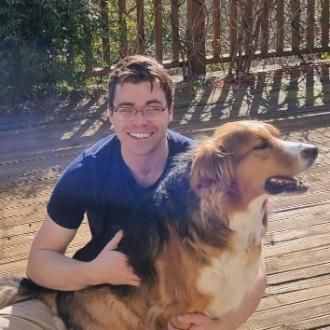 student with short brown hair and glasses smiling. He is petting a brown and white dog